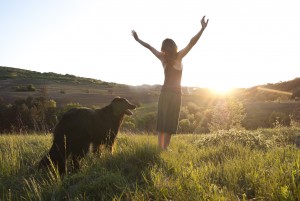 Beautiful young woman raises her arms to the sun whilst walking her dog